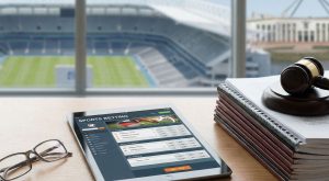 A table with a tablet showing online gambling on the screen and pile of documents where the table is overlooking to a football field